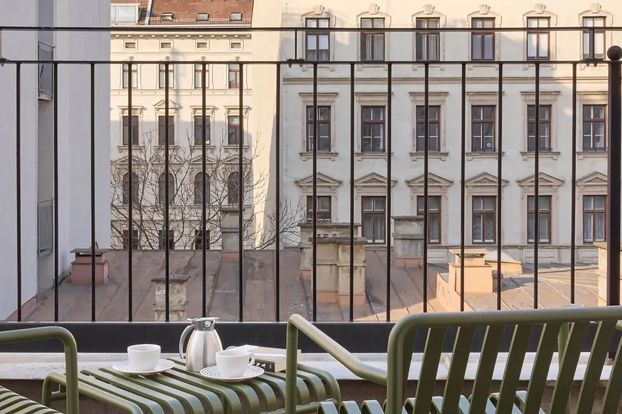 Private Terrasse mit grünen Stühlen, kleinem Tisch und Kaffeegeschirr vor dem Blick auf gegenüberliegende Wiener Altbaufassaden.
