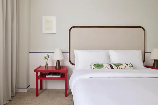 Detail view of a bright hotel room with a large bed, upholstered headboard, red bedside table, and small lamp.