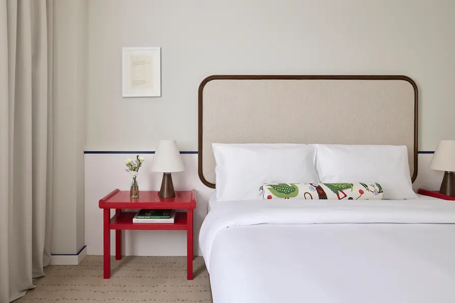 Detailansicht eines hellen Hotelzimmers mit großem Bett, gepolstertem Kopfteil, rotem Nachttisch und kleiner Tischlampe.