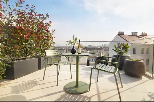 Sunny rooftop terrace with a green table and chairs, a bottle of wine, and a view across the city rooftops.