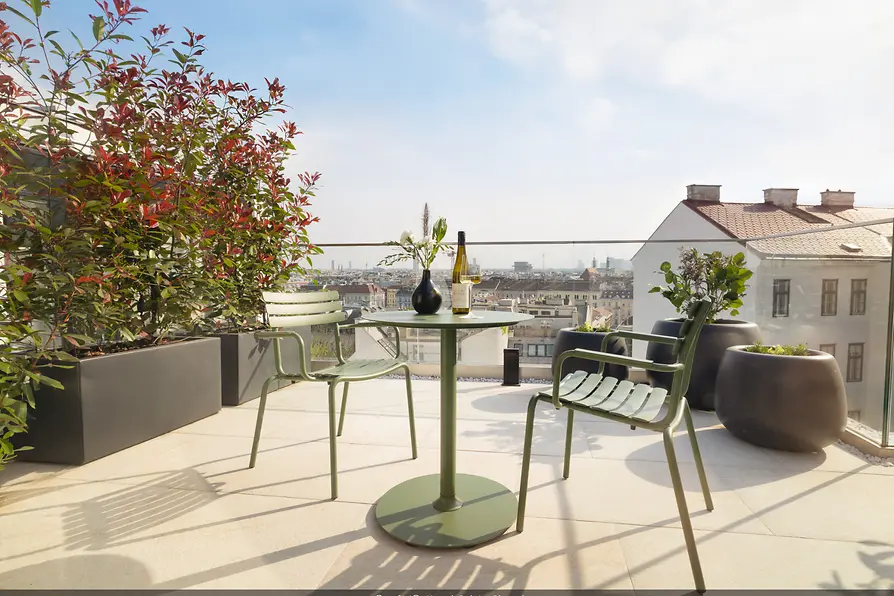 Sunny rooftop terrace with a green table and chairs, a bottle of wine, and a view across the city rooftops.