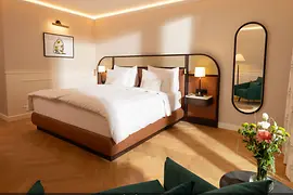 Helles Hotelzimmer mit großem Doppelbett, Fischgrätboden, warmen Beigetönen und Sitzbereich mit grünen Sesseln.