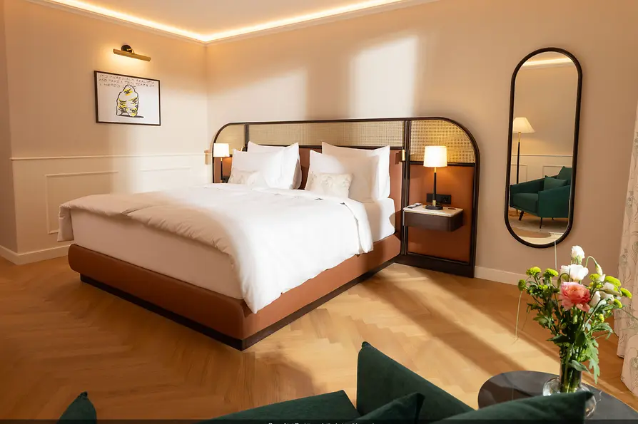 Helles Hotelzimmer mit großem Doppelbett, Fischgrätboden, warmen Beigetönen und Sitzbereich mit grünen Sesseln.