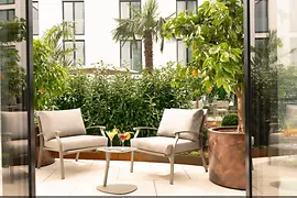 Ruhiger Innenhof mit zwei gepolsterten Sesseln, kleinem Tisch mit Cocktails und üppigen grünen Pflanzen vor heller Fassade.