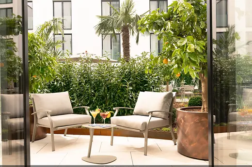Peaceful courtyard with two cushioned armchairs, a small table with cocktails, and lush greenery in front of a light-colored facade.