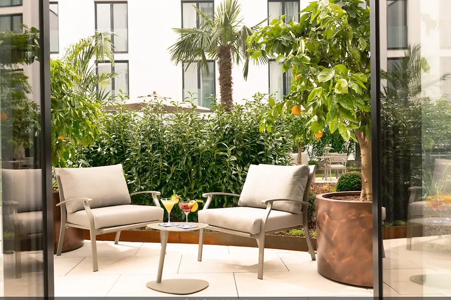 Ruhiger Innenhof mit zwei gepolsterten Sesseln, kleinem Tisch mit Cocktails und üppigen grünen Pflanzen vor heller Fassade.