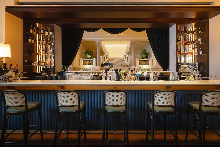 Elegante Bar mit heller Theke, hohen Regalen voller Flaschen und Blick durch dunkle Vorhänge auf eine repräsentative Treppe.