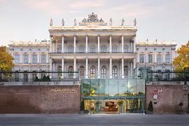 Prächtige historische Fassade des Palais Coburg mit Säulen, Skulpturen und modernem Glaseingang in der Abenddämmerung.
