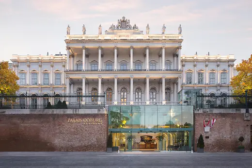 Grand historic facade of Palais Coburg with columns, sculptures, and a modern glass entrance at dusk.