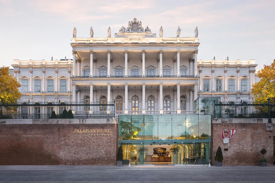 Grand historic facade of Palais Coburg with columns, sculptures, and a modern glass entrance at dusk.