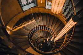 Blick von oben in ein geschwungenes Treppenhaus mit dunklem Geländer und warm leuchtenden Hängelampen im Zentrum.