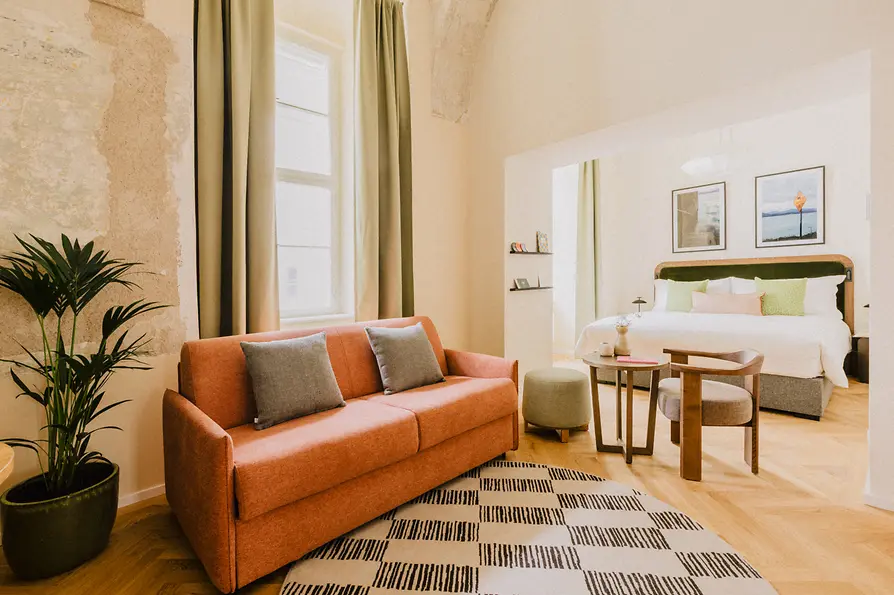 Großzügiges Studio mit rosa Sofa, großem Bett, hellen Vorhängen und kleinem Sitzbereich in sanften Naturtönen.