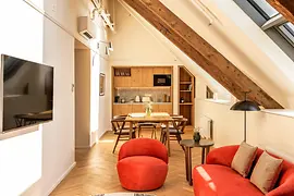 Helles Apartment unter dem Dach mit sichtbarem Holzbalken, Küchenzeile, Esstisch und roten Sesseln im Wohnbereich.