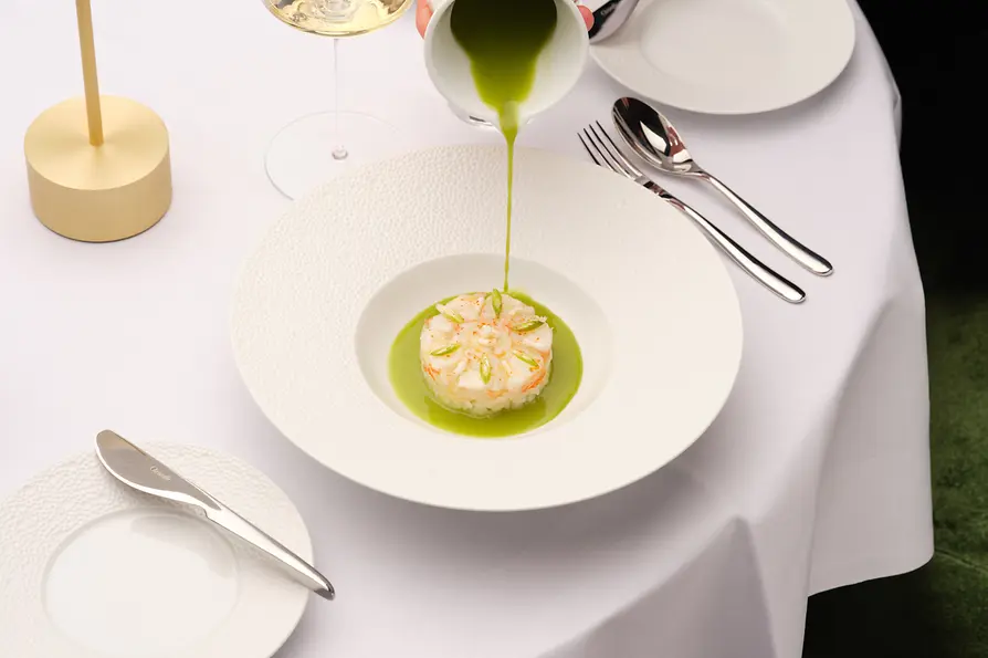 Aus einem kleinen Krug wird leuchtend grüne Sauce über eine fein angerichtete Vorspeise in einem tiefen weißen Teller gegossen.