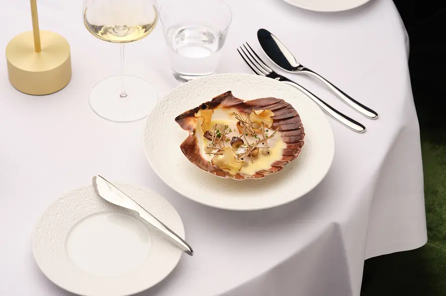 Eine kunstvoll angerichtete Jakobsmuschel liegt in ihrer Schale auf einem weißen Teller, gedeckt auf einer eleganten Tafel mit Wein- und Wasserglas.