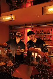 Zwei Bartender arbeiten hinter der Theke; einer gießt einen Drink, der andere bereitet einen Shaker vor, im Hintergrund Spirituosenregale in rotem Licht.
