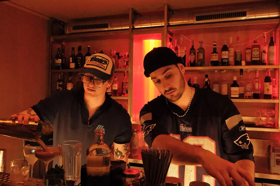 Zwei Bartender arbeiten hinter der Theke; einer gießt einen Drink, der andere bereitet einen Shaker vor, im Hintergrund Spirituosenregale in rotem Licht.