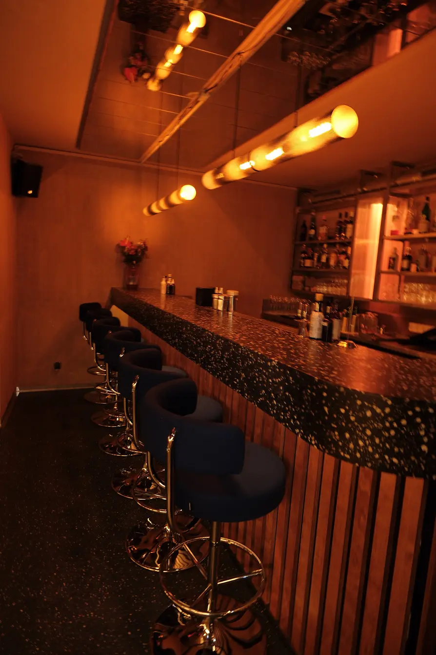 A long terrazzo bar counter with blue bar stools and warm Edison bulbs hanging from the ceiling.