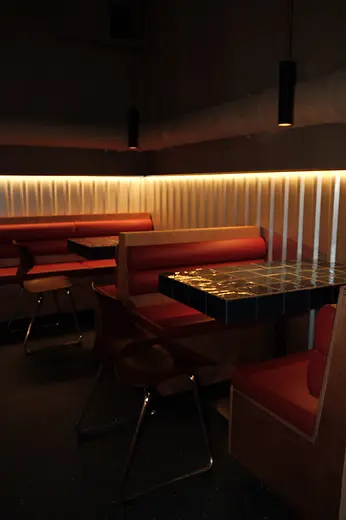 A dimly lit bar area with red upholstered booths, mosaic tile tables, and a warm light strip along the wall.