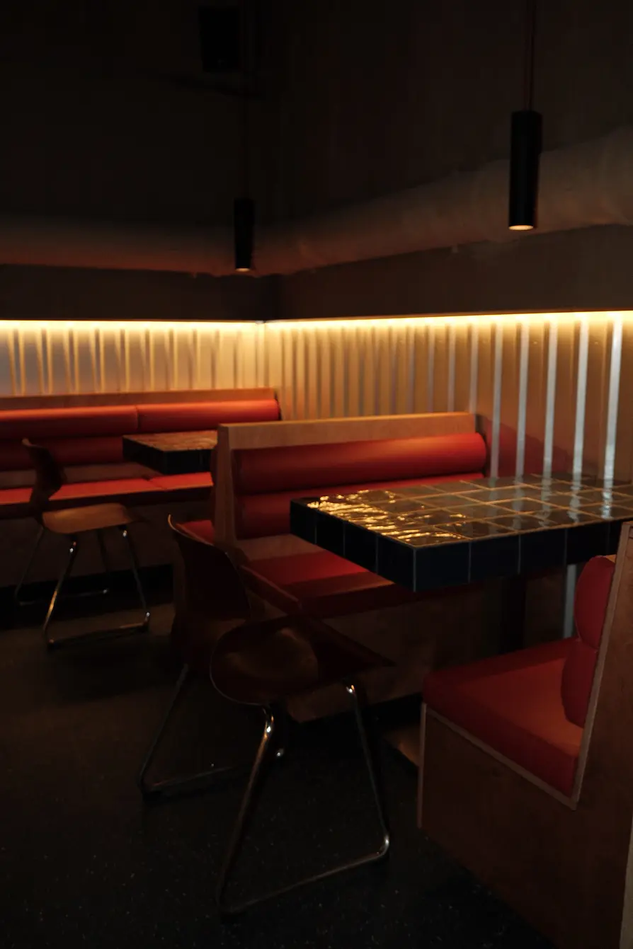 A dimly lit bar area with red upholstered booths, mosaic tile tables, and a warm light strip along the wall.