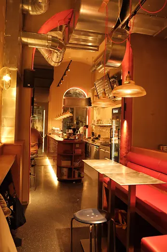 A narrow bar interior with red booth seating, exposed ventilation ducts, and a view of the open kitchen in the background.