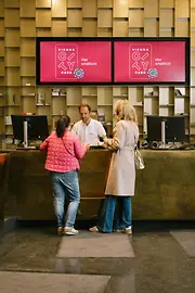 Zwei Besucherinnen sprechen an einem Empfangsschalter der Tourist-Info am Albertinaplatz mit einem Mitarbeiter. Über dem Tresen werben zwei Bildschirme für die Vienna City Card.