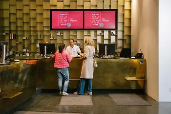 Zwei Besucherinnen sprechen an einem Empfangsschalter der Tourist-Info am Albertinaplatz mit einem Mitarbeiter. Über dem Tresen werben zwei Bildschirme für die Vienna City Card.