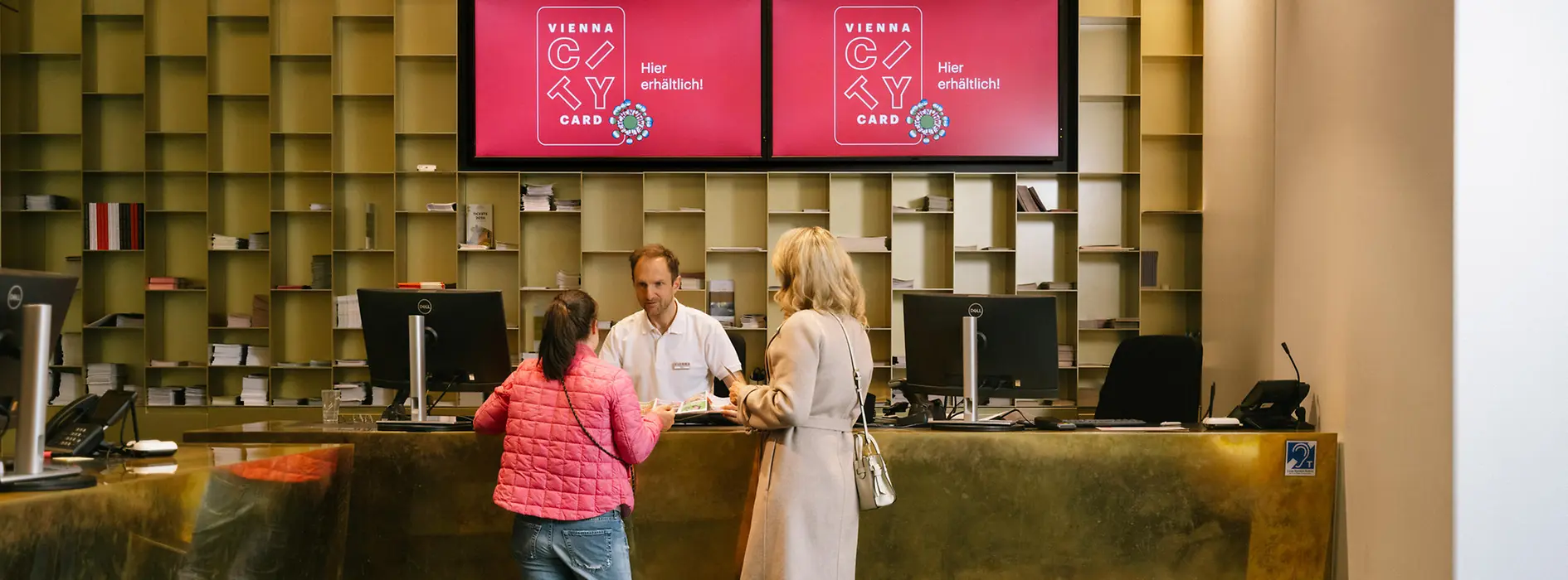 Zwei Besucherinnen sprechen an einem Empfangsschalter der Tourist-Info am Albertinaplatz mit einem Mitarbeiter. Über dem Tresen werben zwei Bildschirme für die Vienna City Card.