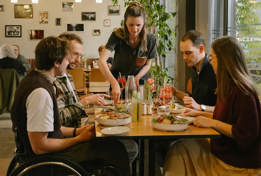 Guests are sitting together at a table at magdas Lokal, including a man using a wheelchair. The image shows an inclusive and accessible restaurant setting.