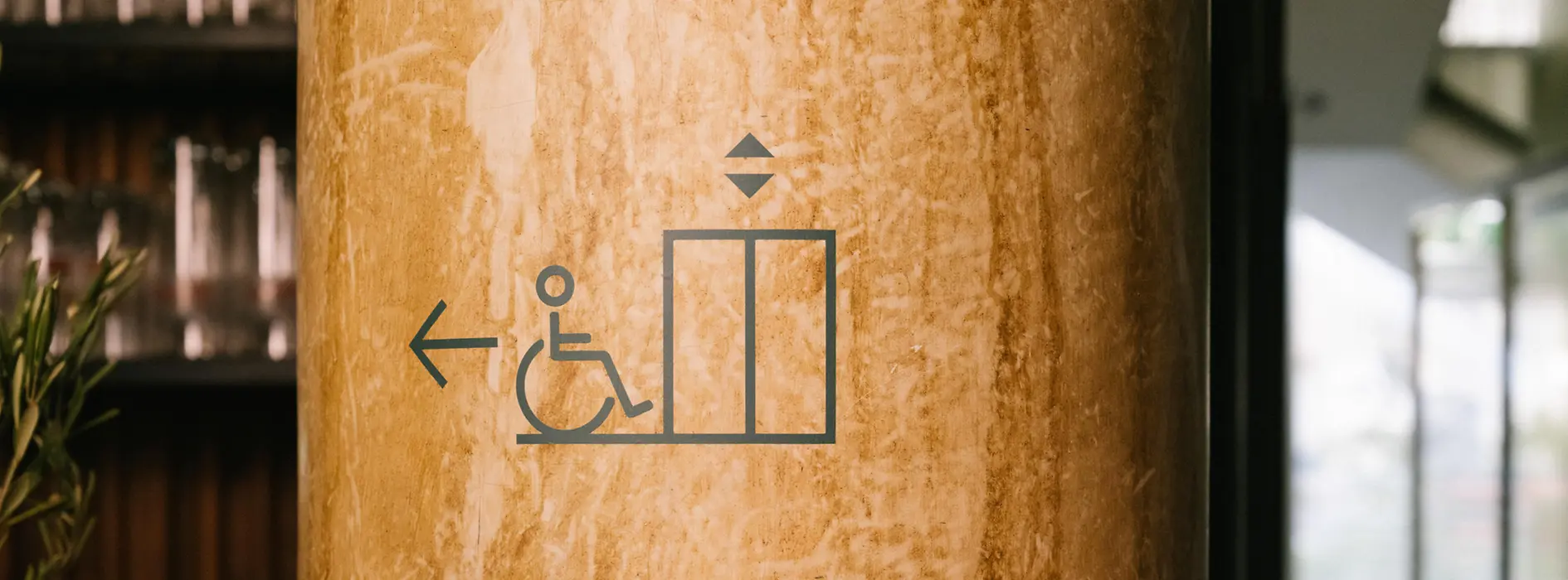 Eine Säule im magdas Lokal zeigt Piktogramme für barrierefreien Zugang mit Lift, WC und Gepäckaufbewahrung. Die Symbole dienen der Orientierung im Innenraum.