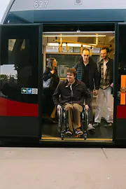 Ein Mann im Rollstuhl fährt aus einer Straßenbahn der Wiener Linien aus, während weitere Fahrgäste im Fahrzeug stehen. Das Bild zeigt barrierefreie Mobilität im öffentlichen Verkehr.