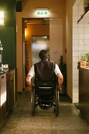 Ein Mann im Rollstuhl fährt durch den Innenbereich des magdas Lokal in Richtung Aufzug. Das Bild zeigt barrierefreie Wegeführung im Lokal.
