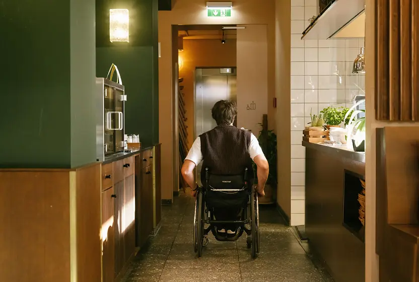 Ein Mann im Rollstuhl fährt durch den Innenbereich des magdas Lokal in Richtung Aufzug. Das Bild zeigt barrierefreie Wegeführung im Lokal.