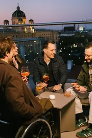 Mehrere Personen sitzen gemeinsam auf der Terrasse der MQ Libelle, darunter ein Mann im Rollstuhl, mit Blick über die Stadt. 