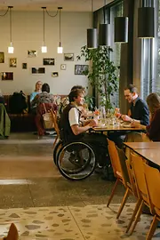 Ein Mann im Rollstuhl befindet sich im Gastraum des magdas Lokal und ist Teil einer Gruppe am Tisch. Das Bild zeigt barrierefreie Teilhabe im Restaurantalltag.