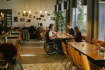 Ein Mann im Rollstuhl befindet sich im Gastraum des magdas Lokal und ist Teil einer Gruppe am Tisch. Das Bild zeigt barrierefreie Teilhabe im Restaurantalltag.