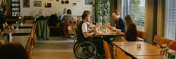 Ein Mann im Rollstuhl befindet sich im Gastraum des magdas Lokal und ist Teil einer Gruppe am Tisch. Das Bild zeigt barrierefreie Teilhabe im Restaurantalltag.