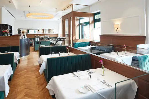 Interior of the Plachutta flagship restaurant with set tables, dark green banquettes and a member of staff.