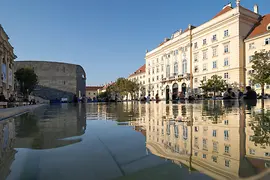 Flache Wasserfläche im MuseumsQuartier spiegelt eine helle historische Fassade und das dunkle mumok-Gebäude unter blauem Himmel.