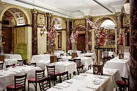 Prunkvoller Speisesaal mit Marmorsäulen, Rundbögen, hohen Blumenarrangements und weiß gedeckten Tischen in klassischem Ambiente.