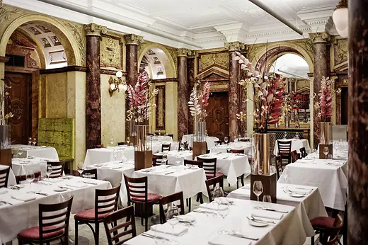 Grand dining room with marble columns, arches, tall floral arrangements, and white tablecloth tables in a classic setting.