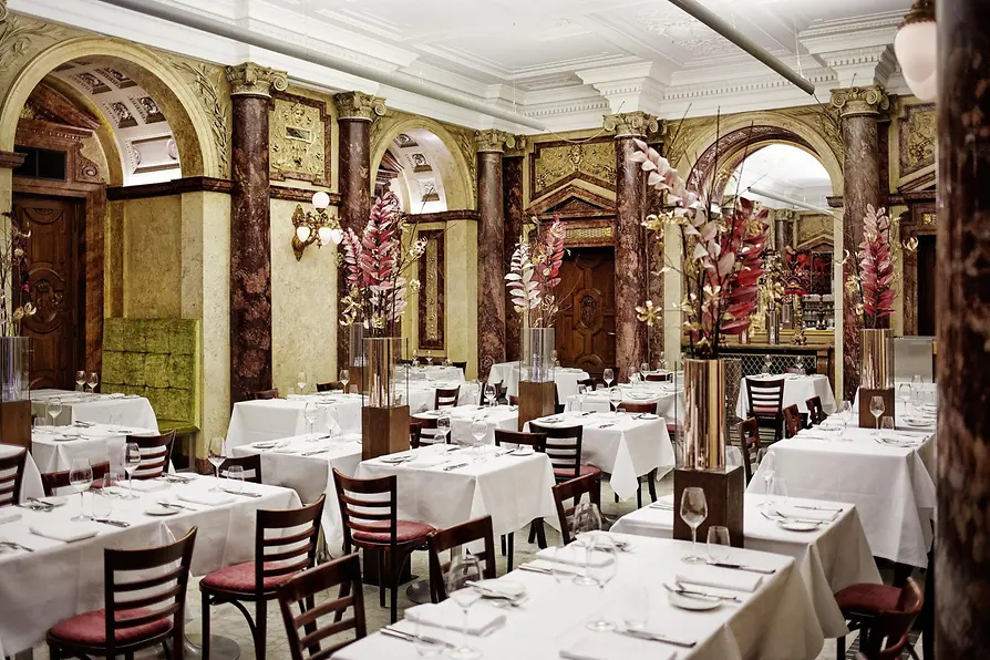 Grand dining room with marble columns, arches, tall floral arrangements, and white tablecloth tables in a classic setting.