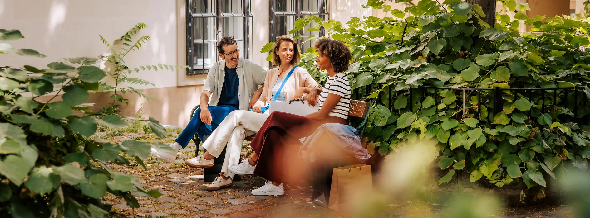 Two women and a man on a bench in a leafy courtyard