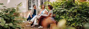 Two women and a man on a bench in a leafy courtyard