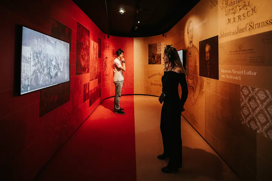 Two visitors in an exhibition space designed in yellow and red in front of screens.