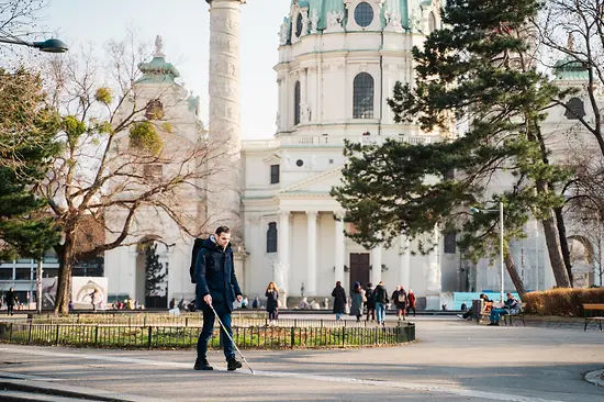 Blinde Person geht mit einem Blindenstock vor der Wiener Karlskirche am Blindenleitsystem entlang